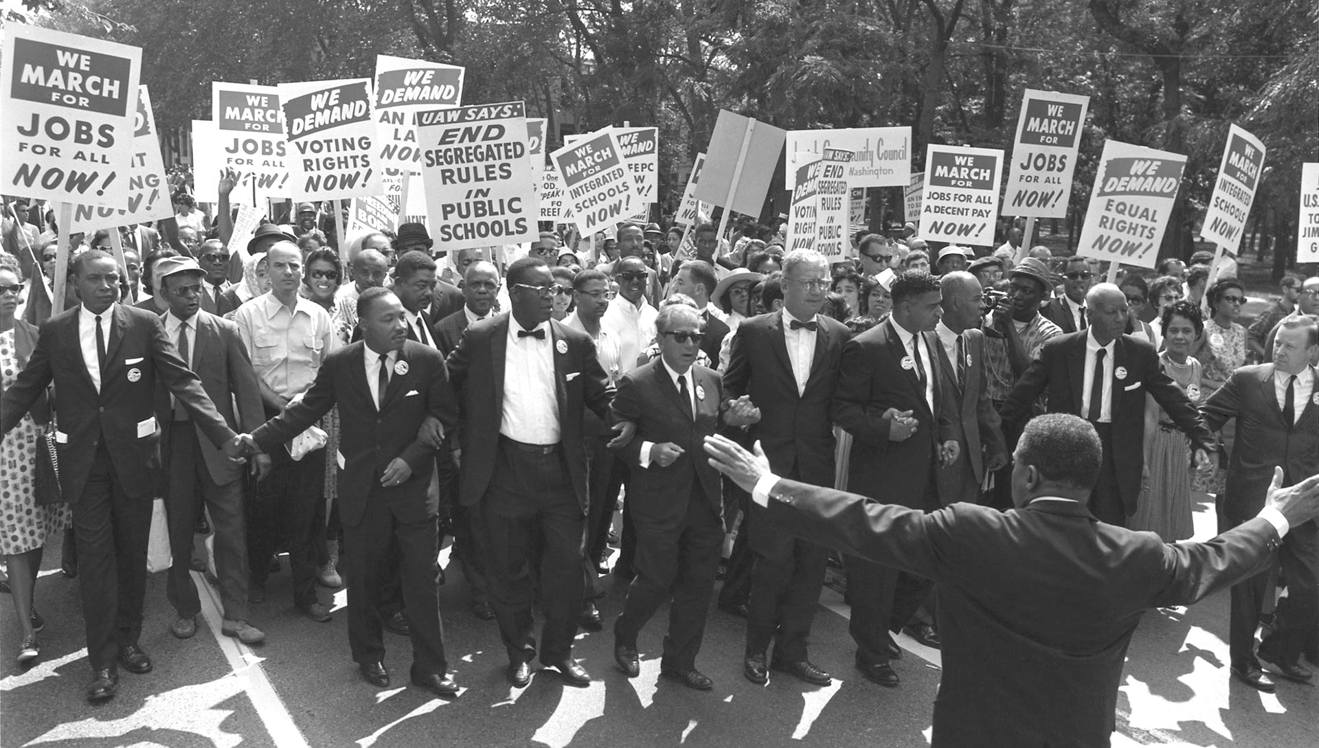 A historic black-and-white photo showing a crowd of people marching with signs advocating for jobs and civil rights. The group, which includes some individuals in suits, is walking arm in arm on a tree-lined street.